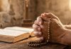 Imagem de mãos em oração segurando um terço, simbolizando a gratidão e a força na vida cristã sob uma luz suave de manhã.